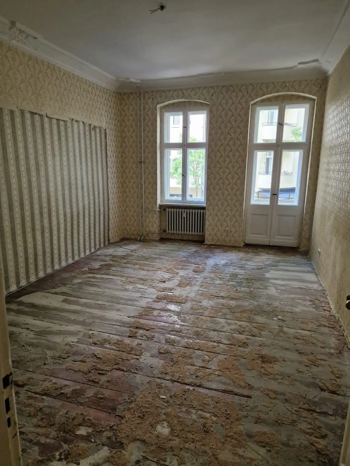 Zimmer mit Balkontür vor der Sanierung – alte Tapete und beschädigter Dielenboden
